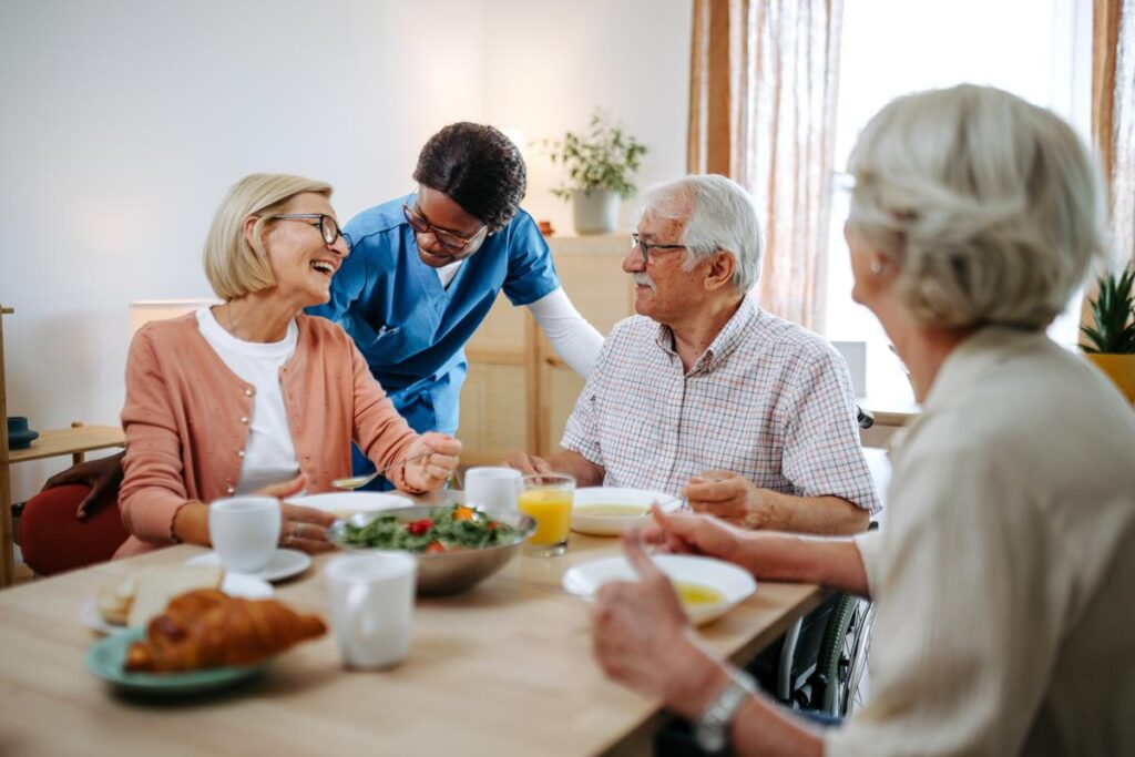 A smiling nurse checks on senior residents during lunch at a nursing home. She engages in friendly conversation with the elderly adults as they enjoy their meal.